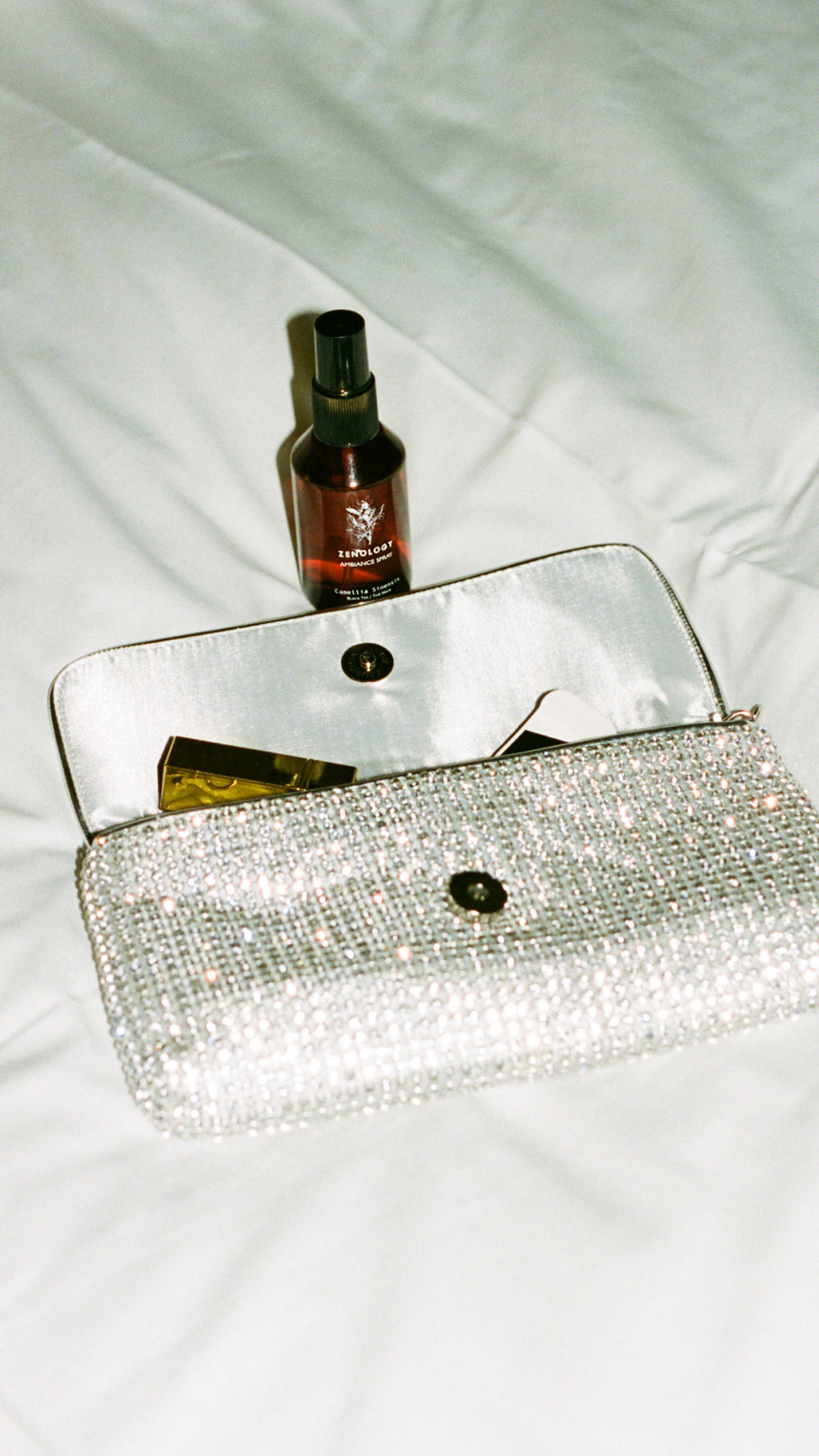 A shimmering, white beaded clutch purse is open on a bed with white sheets. Partially visible items inside include a lipstick and a compact mirror. Behind the clutch, a small brown ZENOLOGY Ambiance Travel Spray Camellia Sinensis bottle is placed upright.