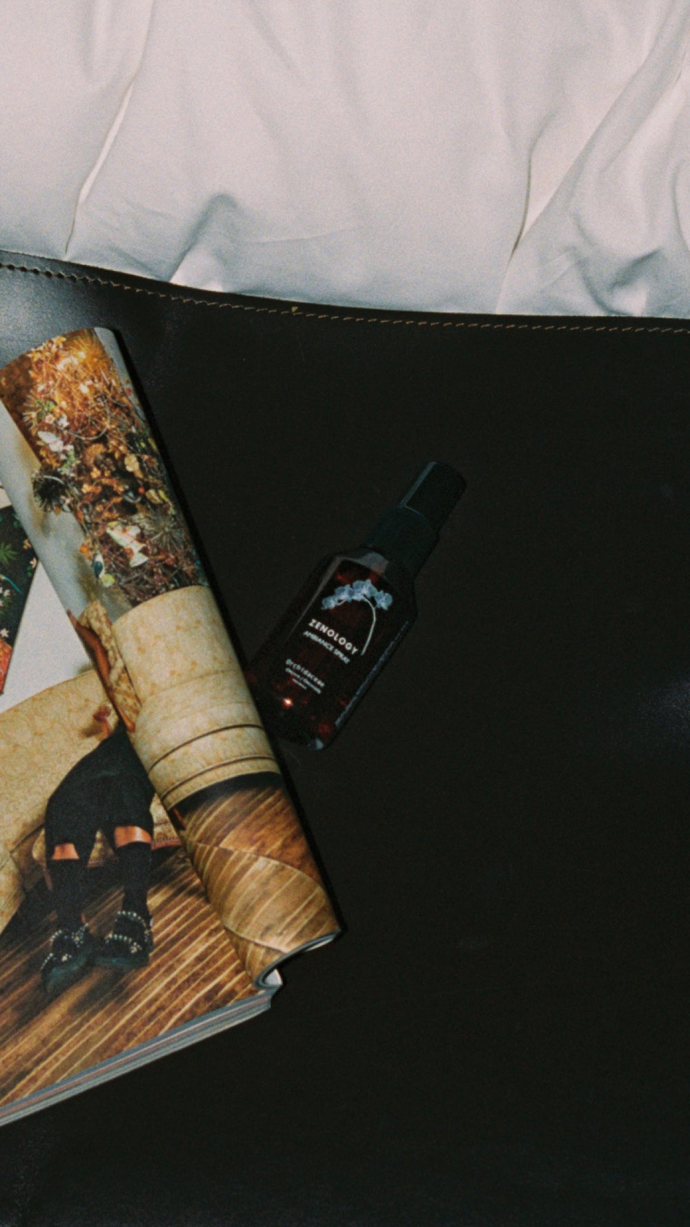 A spray bottle of ZENOLOGY Ambiance Travel Spray Orchidaceae sits on a dark surface next to an open magazine with colorful photos. The top edge of a white pillow is visible at the top of the image.