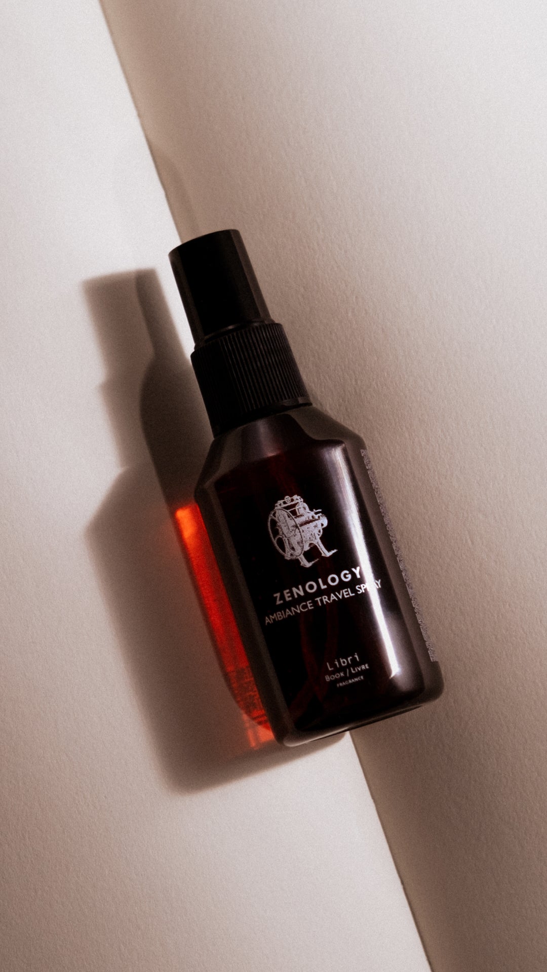 A dark-colored bottle of ZENOLOGY Ambiance Travel Spray labeled "Libri" lies on a light surface, casting a distinct shadow. The bottle features a pump spray mechanism and white text with a small graphic. Light and shadow create a dramatic effect.