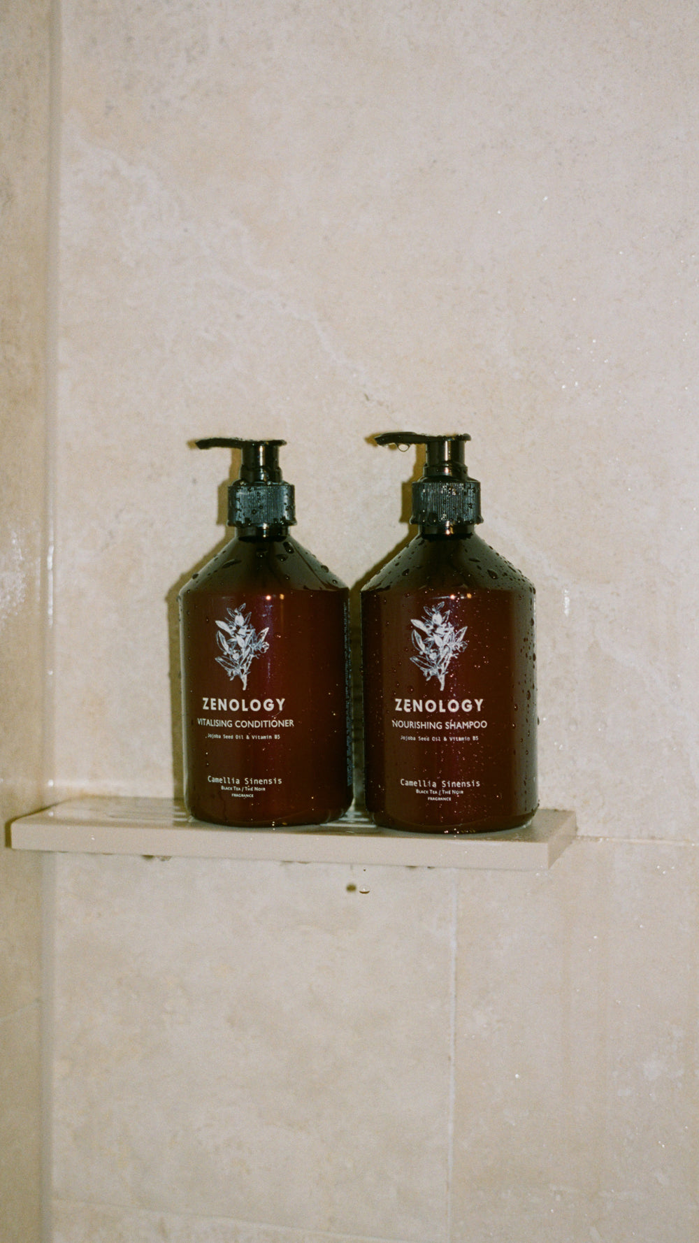 Two bottles of ZENOLOGY Camellia Sisensis hair care products are placed on a shower shelf with a beige tile background. The bottle on the left is a "Nourishing Conditioner," and the one on the right is a "Nourishing Shampoo." Both bottles feature a pump dispenser and a minimalist design.