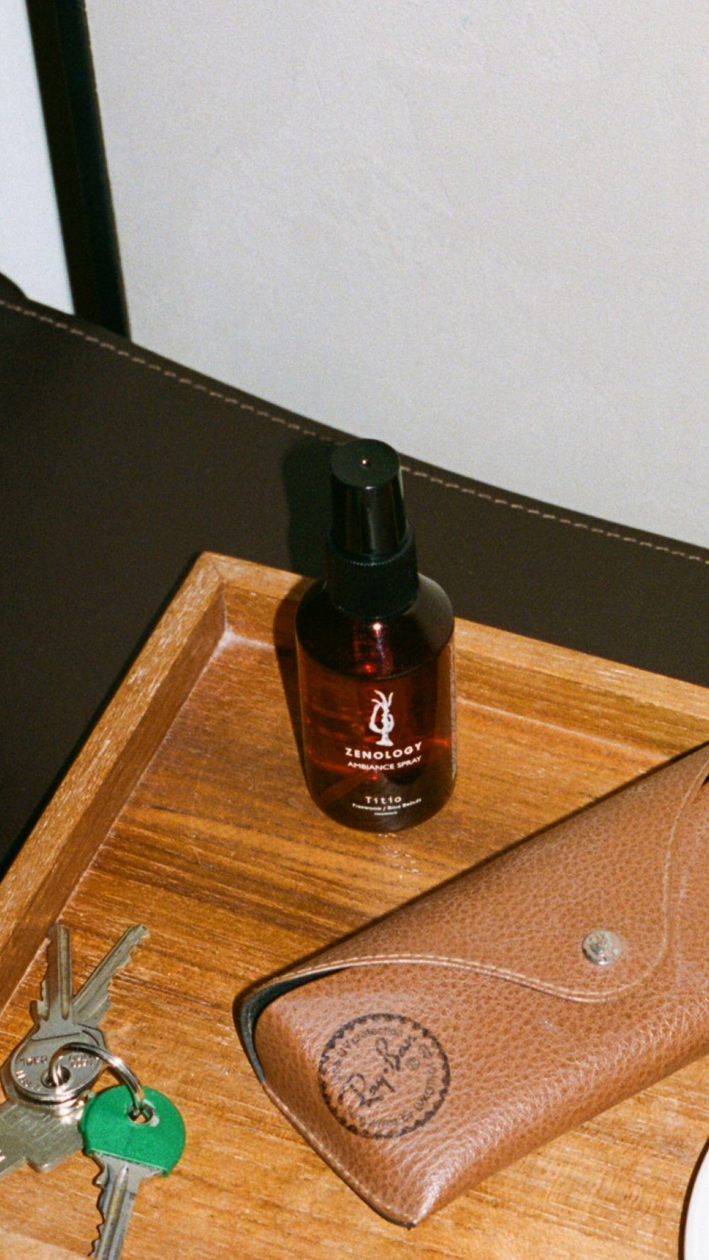 A wooden tray holds a brown spray bottle labeled "ZENOLOGY Ambiance Travel Spray Titio" and a brown leather case with a button clasp. A set of keys with a green tag rests beside the tray, on a dark surface.