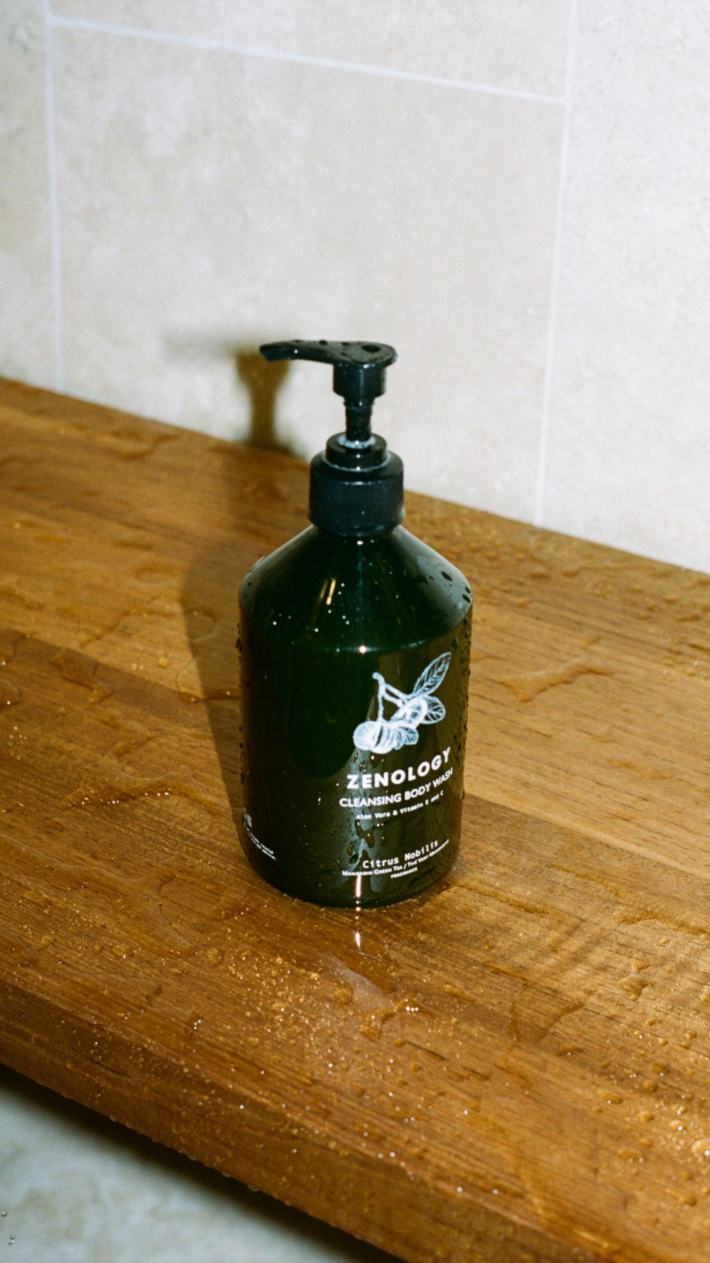 A green bottle of ZENOLOGY Citrus Nobilis Cleansing Body Wash with a black pump dispenser is placed on a wet wooden surface. The background features light-colored tiles. Water droplets are visible on the bottle and the surface.