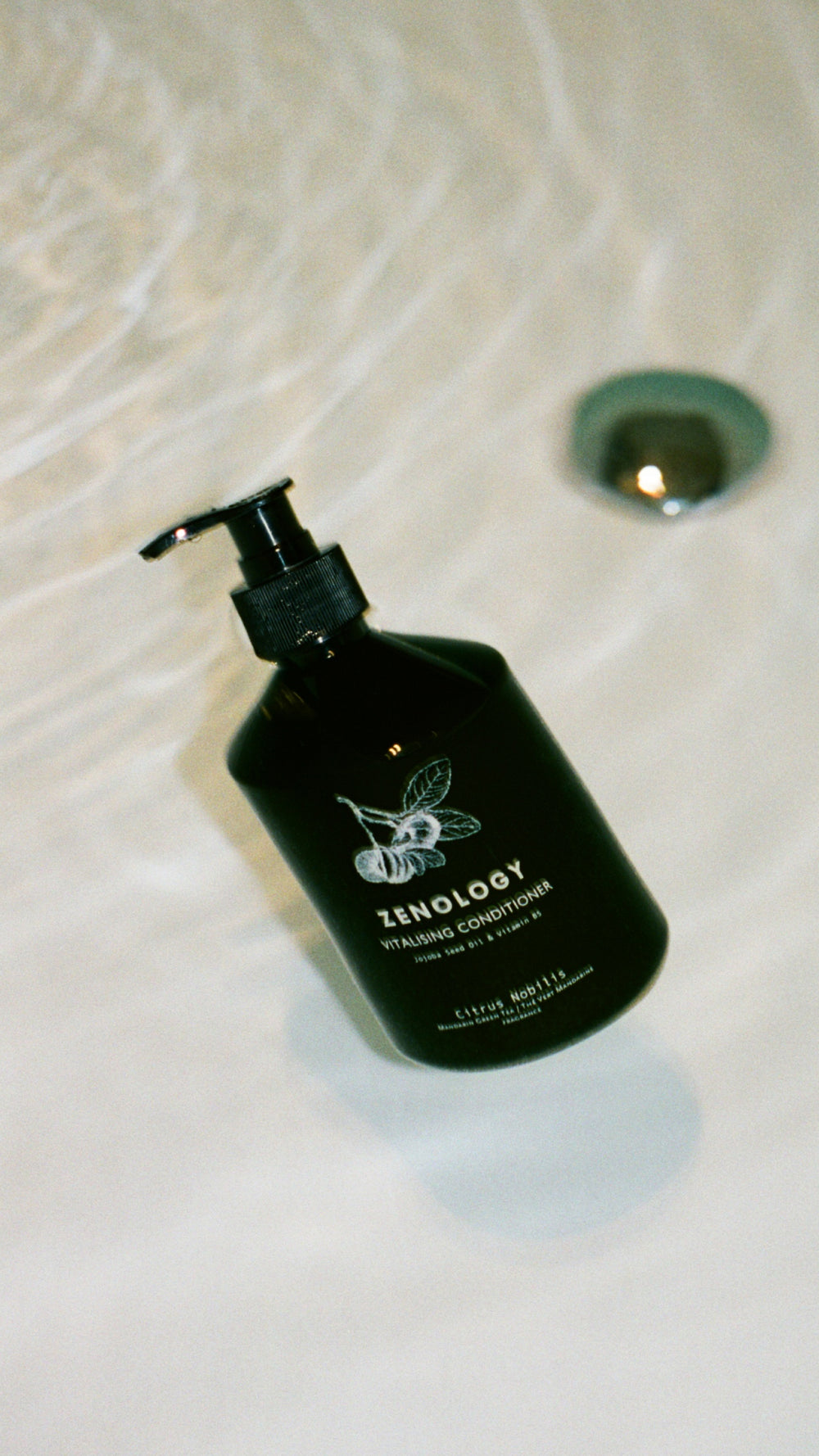 A green bottle of ZENOLOGY Vitalizing Conditioner with a pump dispenser floating in a white bath tub filled with water. Water is flowing into the tub from the top of the image, creating ripples around the bottle.