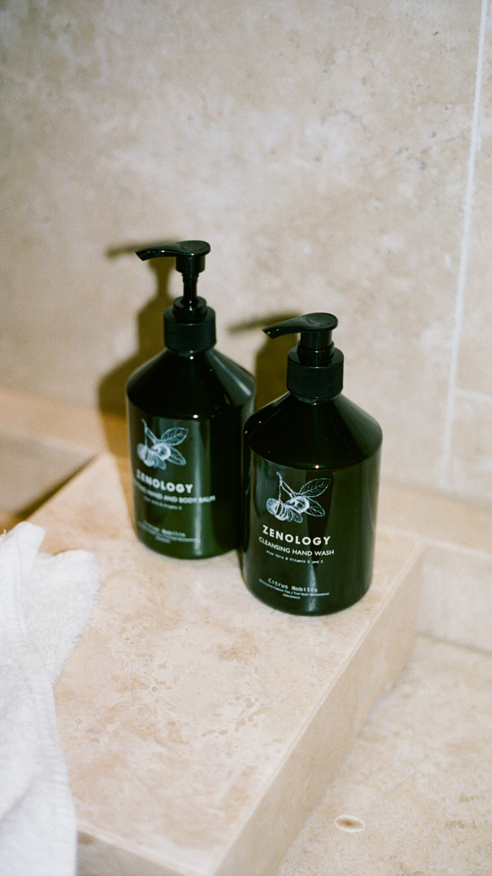 Two bottles of ZENOLOGY Citrus Nobilis with pump dispensers placed on a beige stone surface, near a white towel. The bottles are green with minimalist labels featuring text and a leaf design. One is labeled Cleansing Hand Wash and the other Hydrating Hand and Body Balm. The background includes beige tiles, suggesting a bathroom setting.