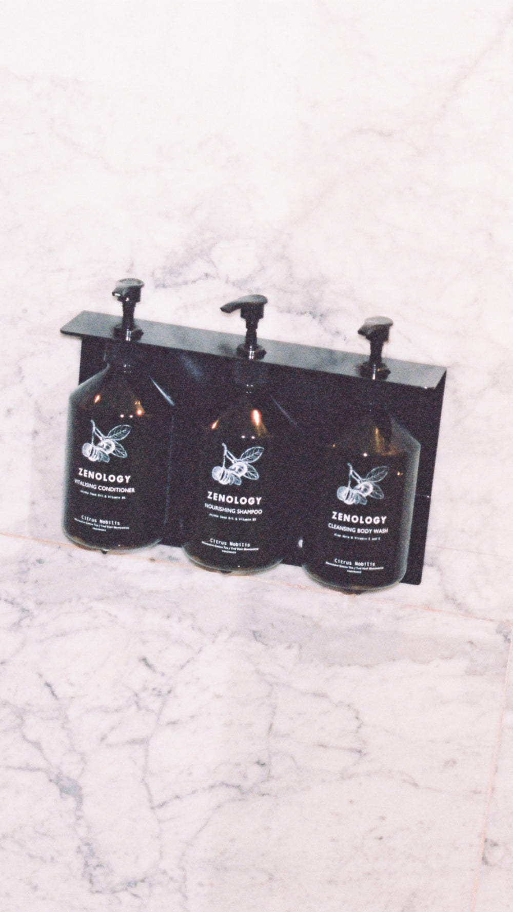 Three dark-green colored ZENOLOGY bottle dispensers are mounted on a marble wall. Each bottle features a white label with Citrus Nobilis branding and text, filled with different specified toiletries for use.
