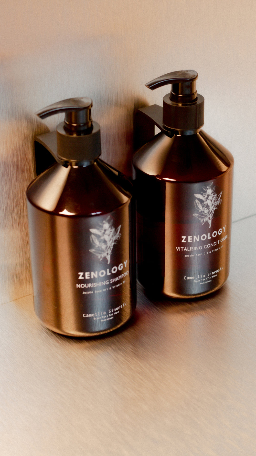 Two brown pump bottles labeled "ZENOLOGY" are mounted on a metal wall. The left bottle reads "Nourishing Shampoo" and the right bottle reads "Vitalising Conditioner." Both bottles display botanical illustrations and the subtitle "Camellia Sinensis.