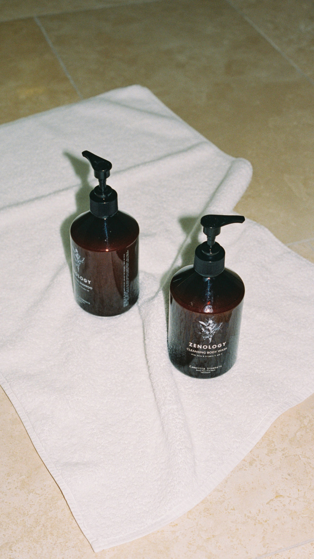 Two brown ZENOLOGY Camellia Sinensis pump bottles are placed on a white towel spread out on a beige tiled floor. Both bottles appear to be identical in size, design, and label.
