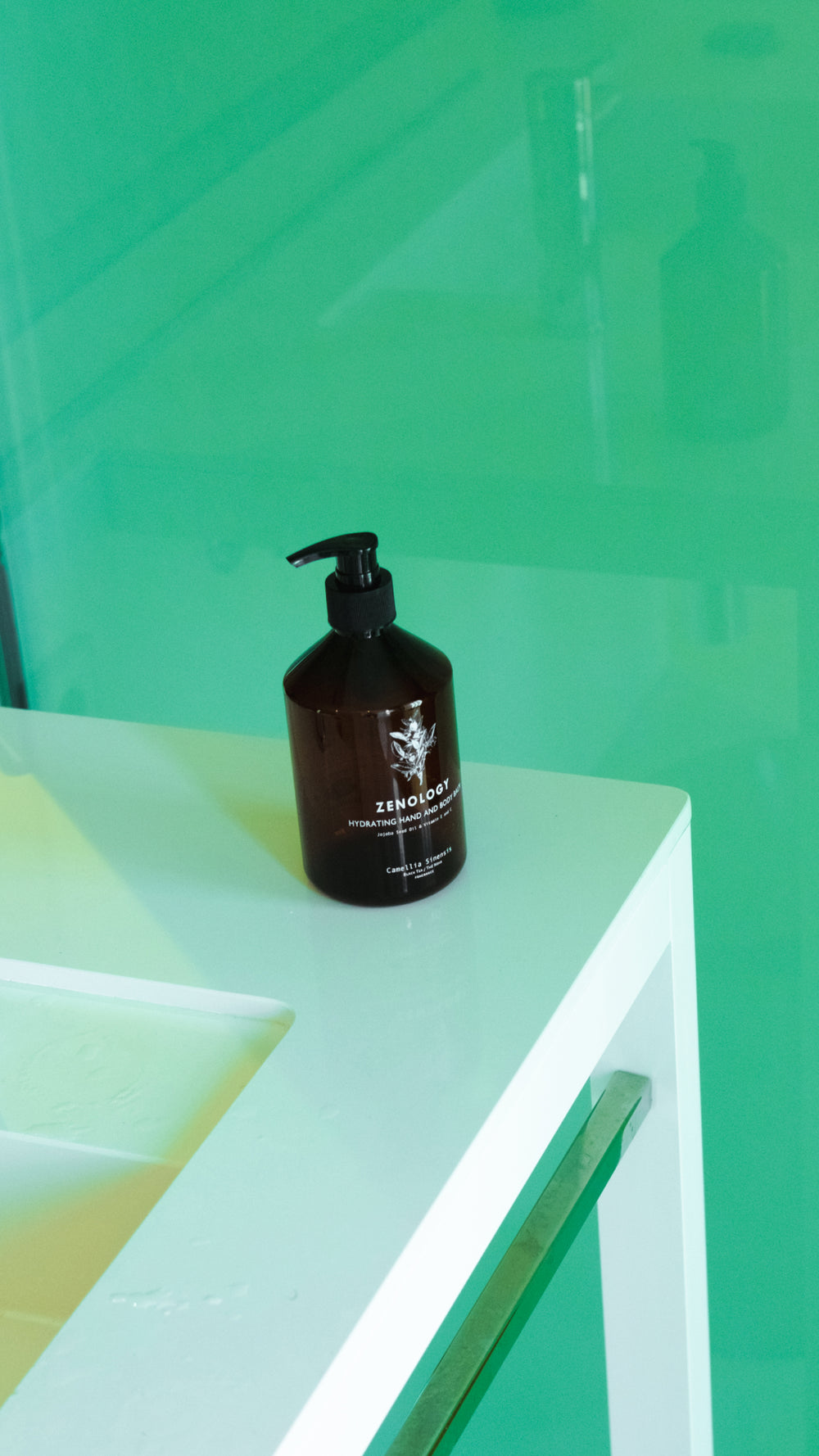 A brown ZENOLOGY bottle of Camellia Sinensis Hand and Body Balm with a black pump top sits on the edge of a white sink in a modern bathroom with a green translucent glass wall. Reflections of the bottle and the sink can be seen on the glass surface.