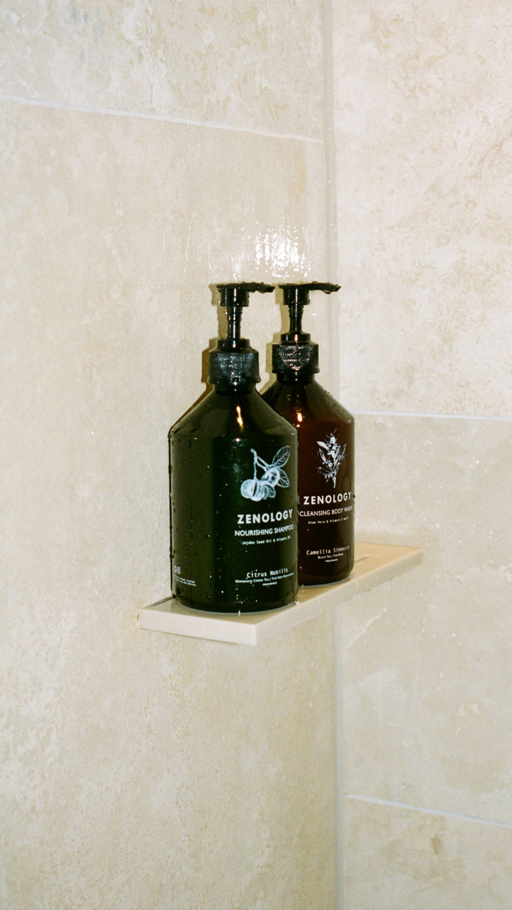 Two ZENOLOGY bottles, one labeled Citrus Nobilis Nourishing Shampoo and the other Camellia Sinensis Cleansing Body Wash, sit on a small shower shelf against a beige tiled wall. Both bottles have pump dispensers and are slightly wet from recent use.