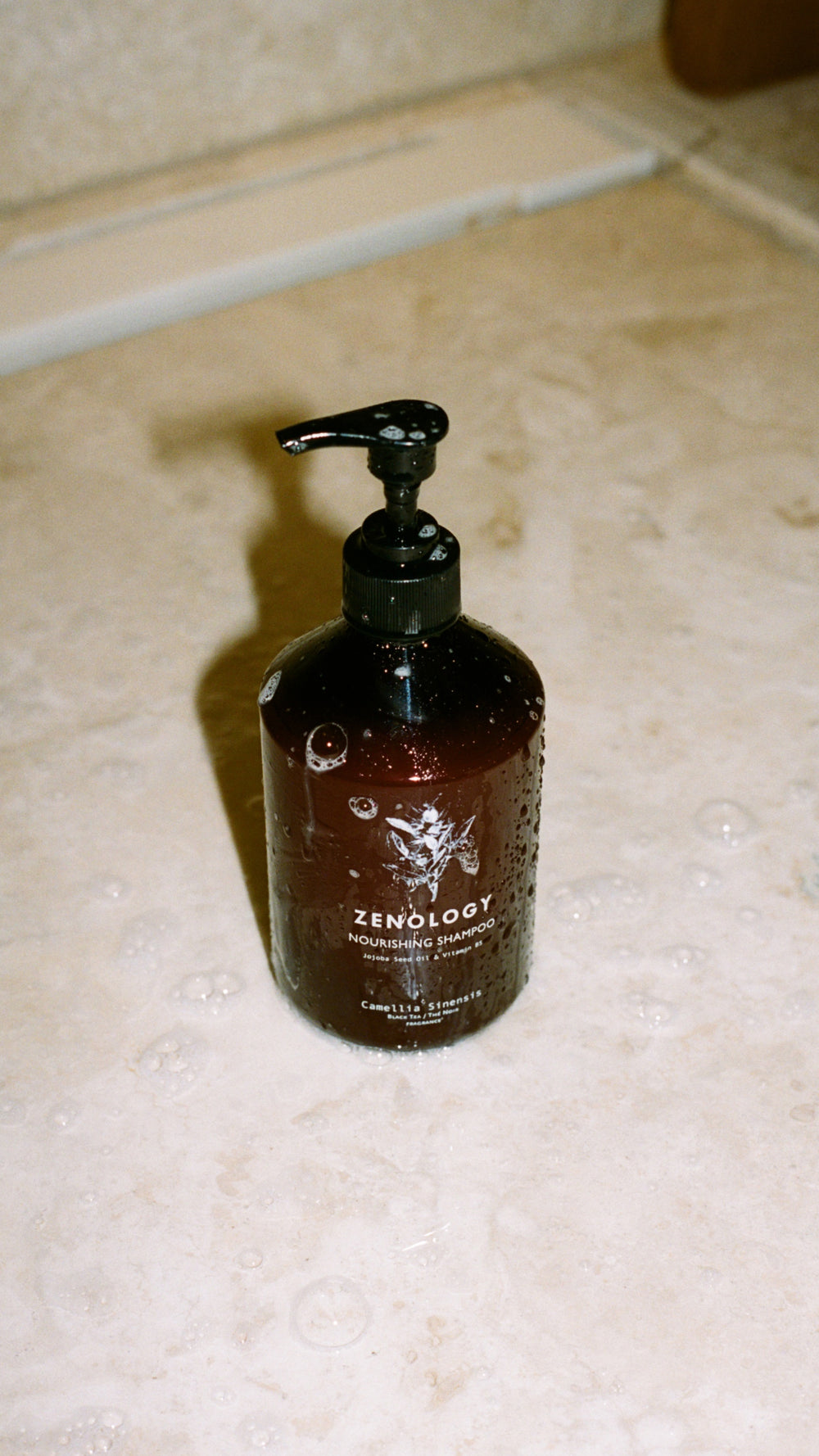 A brown bottle of ZENOLOGY Camellia Sinensis Nourishing Shampoo with a black pump dispenser is placed on a wet, beige tiled surface. The bottle has some drops of water on it. The background shows a slightly blurred edge of a bathtub or sink.