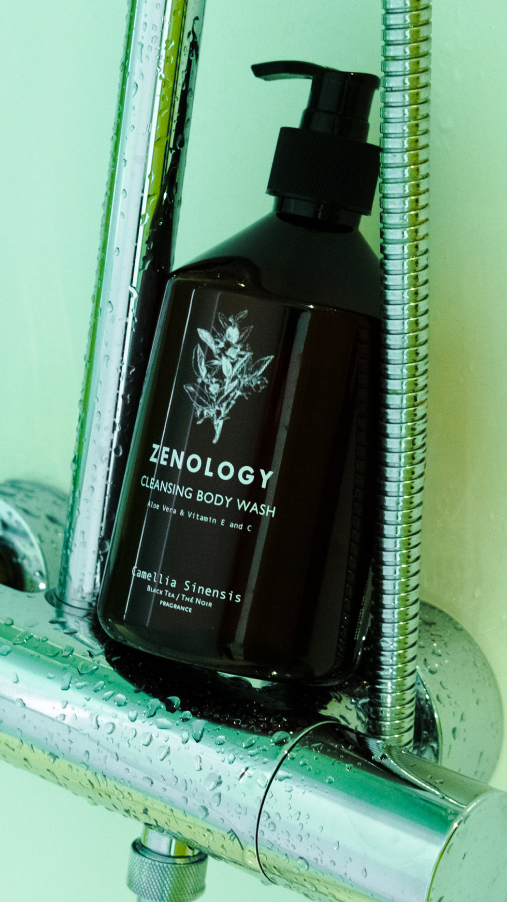 A bottle of ZENOLOGY Cleansing Body Wash with a pump dispenser is placed on a metal shower rack. The label on the dark bottle highlights active ingredients such as aloe vera, vitamin E, and Camellia Sinensis. Water droplets are visible on the metal rack and bottle.