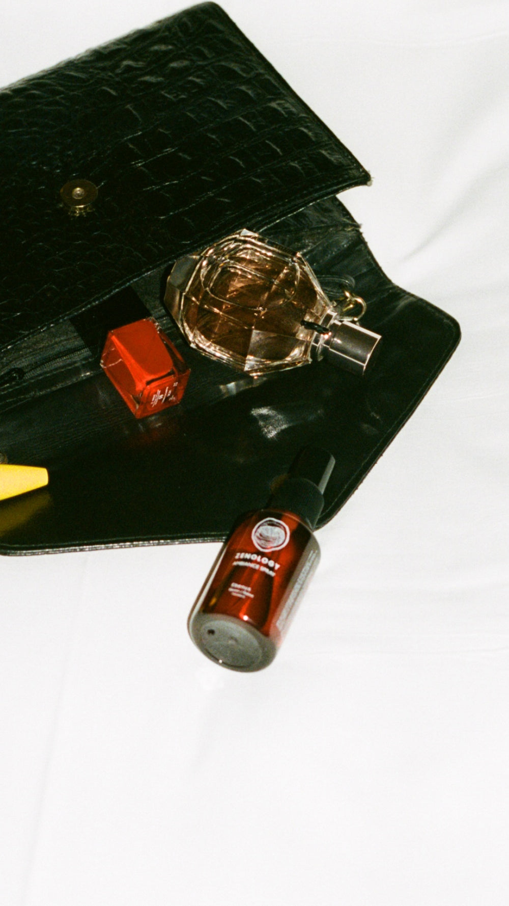 An open black clutch is on a white surface, revealing its contents: a bottle of nail polish, a small perfume bottle shaped like a gem, and a brown bottle of  ZENOLOGY Ambiance Travel Spray in Ebenus. A yellow item is partially visible at the edge of the clutch.