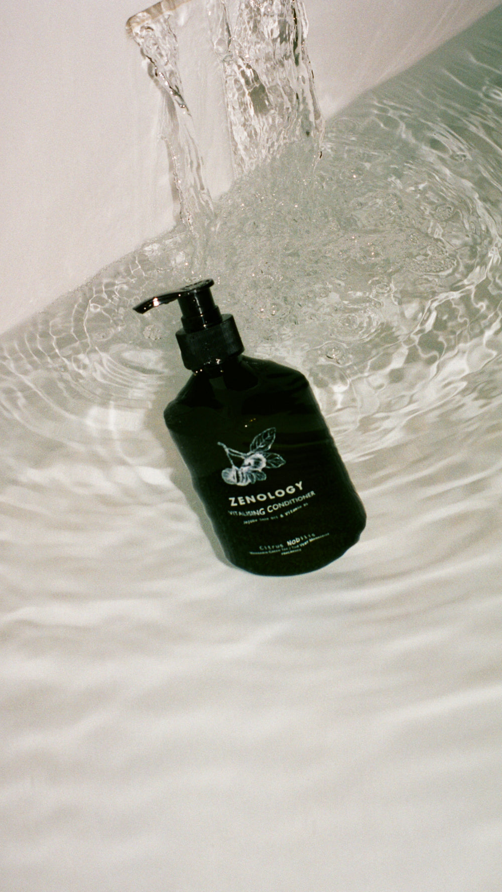A green bottle of ZENOLOGY Citrus Nobilis Vitalizing Conditioner with a pump dispenser floating in a white bath tub filled with water. Water is flowing into the tub from the top of the image, creating ripples around the bottle.