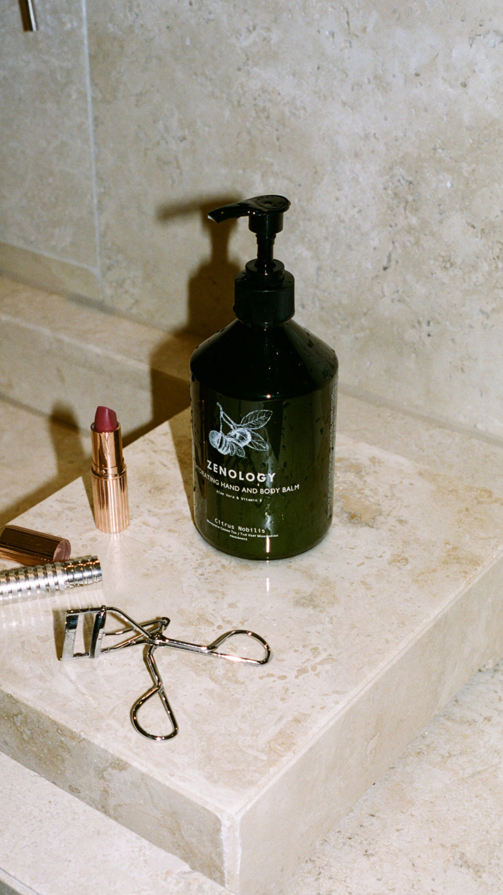 A green bottle of ZENOLOGY Citrus Nobilis Hand and Body Balm sits on a beige marble surface. Surrounding the bottle are a tube of lipstick with the cap off, a closed lipstick tube, and an eyelash curler. The background features a beige tiled wall.