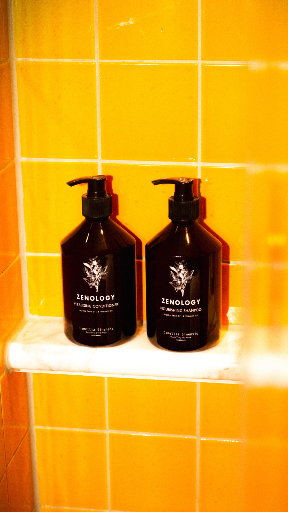 Two brown bottles of ZENOLOGY hair products, one Vitalizing Conditioner and one Nourishing Shampoo, are placed on a white shower shelf. The background is composed of bright, yellow-orange tiled walls, giving the setting a warm ambiance.