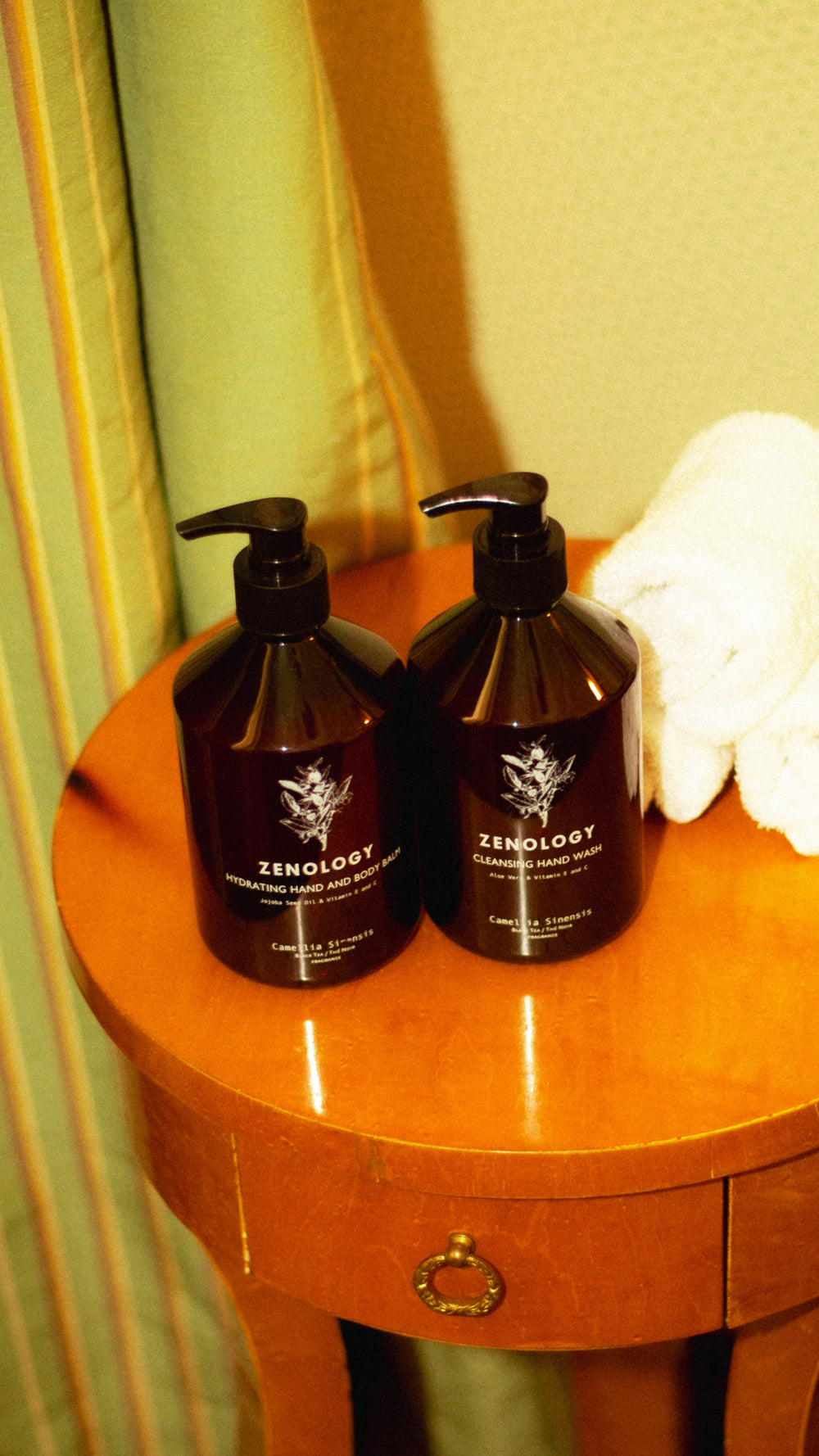 Two brown pump bottles of ZENOLOGY Camellia Sinensis Hydrating Hand and Body Balm and Cleansing Hand Wash sit on a small wooden side table, next to a neatly rolled white towel. The labels feature a botanical design, and the background includes striped curtains.