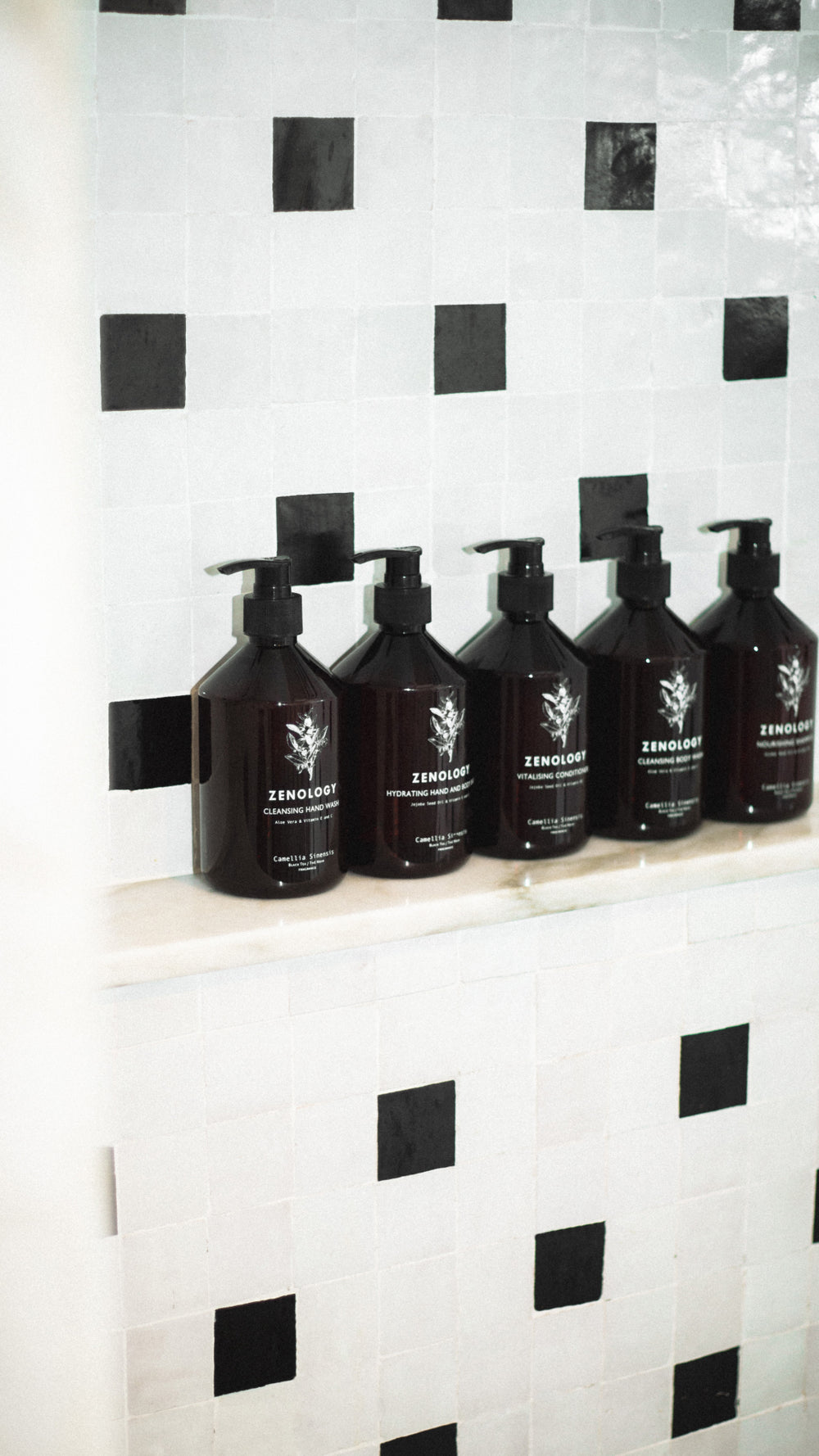 Five dark brown bottles of ZENOLOGY Camellia Sinensis arranged in a row on a tiled shelf. The shelf is part of a wall decorated with black and white square tiles. Each bottle has a black pump top and the labels feature a minimalist design with an illustrative logo