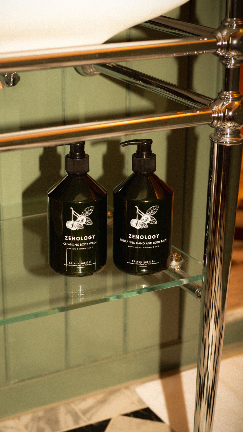 Two green bottles of ZENOLOGY Citrus Nobilis rest on a glass shelf supported by a metallic frame. One is labeled Cleansing Body Wash and the other Hydrating Hand and Body Balm. The background features a green paneled wall and part of a patterned floor.