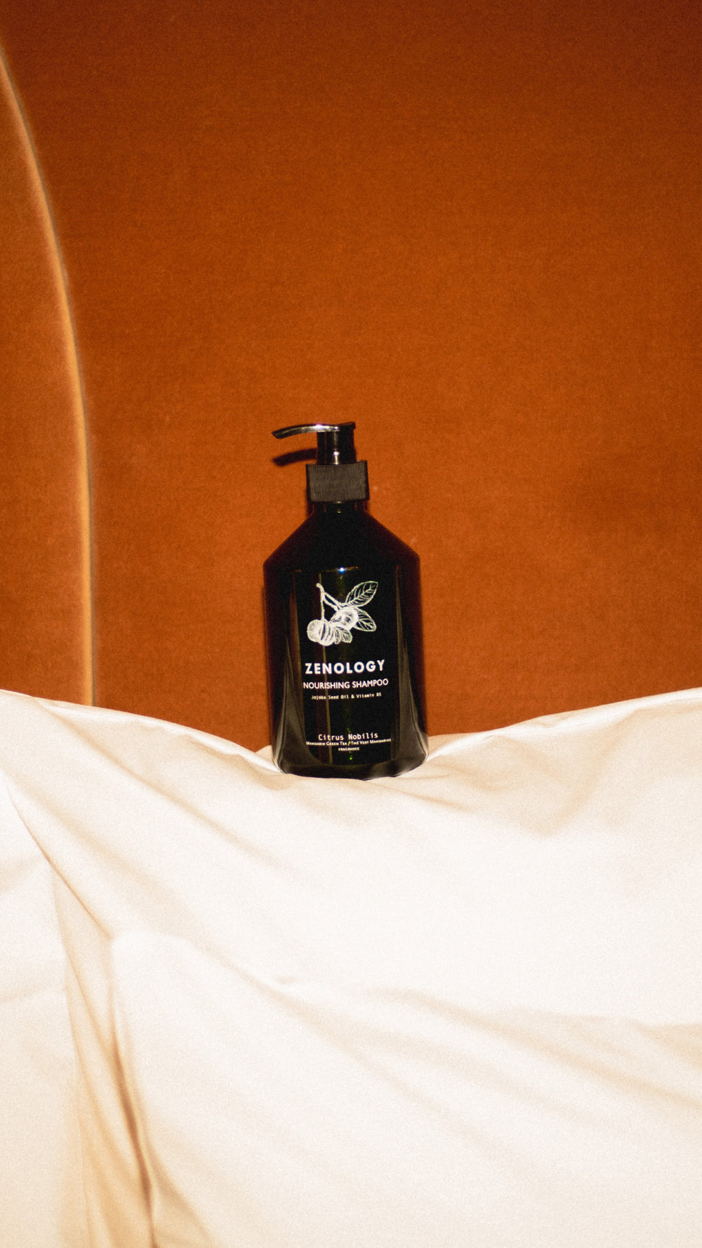 A green pump bottle labeled ZENOLOGY Citrus Nobilis rests against white pillows and a brown fabric background. The unique bottle design and elegant label suggest a premium product, possibly a cosmetic or skincare item.